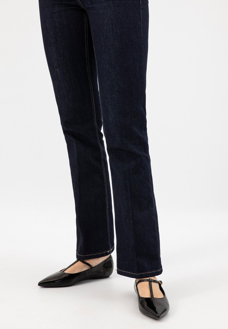 Gambe con jeans blu scuro a gamba dritta e ballerine nere con cinturino a T e punta appuntita su sfondo bianco.