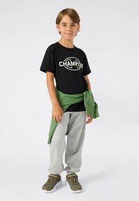 Svart T-skjorte med "CHAMPION"-logo, grå joggebukser, grønn genser knyttet rundt livet, og grønne sko med hvite såler.