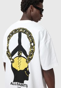 Hvid oversized t-shirt med sort og gul grafik af et stiliseret hoved med et fredssymbol og kædedesign på ryggen.