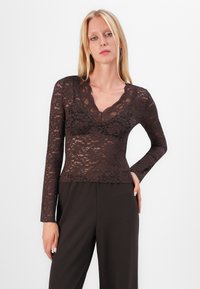 Langermet brun blondetopp med bølgete V-hals og blomstermønster, paret med mørke bukser. Stoffet er gjennomsiktig og strukturert.