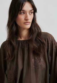 Blouse transparente marron avec broderie florale en marron plus foncé. Caractéristiques incluent un col rond et une silhouette ample et fluide.
