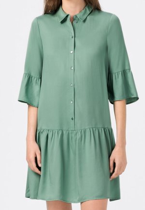 Frau trägt ein locker sitzendes, mintgrünes Kleid mit Knöpfen, gerafften dreiviertel Ärmeln und einem gerafften Rocksaum.