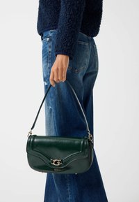 Borsa in pelle verde scuro con design curvato, dettagli in metallo dorato e tracolla removibile, tenuta da una persona che indossa jeans blu.