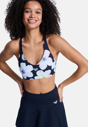 Uśmiechnięta kobieta o kręconych włosach, ubrana w czarny sportowy biustonosz w kwiaty i dopasowaną czarną sportową spódnicę, z rękami opartymi na biodrach.