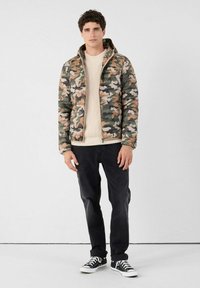 Camouflage donsjack met capuchon, beige gebreide trui, zwarte broek en zwarte sneakers met witte veters. Gestructureerde patronen zichtbaar op het jack.
