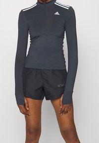 Donna che indossa una maglia da atletica nera a maniche lunghe Adidas con strisce bianche e pantaloni corti neri Adidas su uno sfondo semplice.