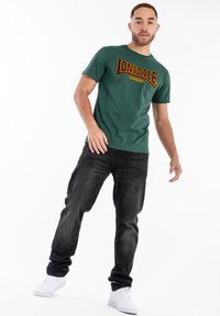 Grøn bomulds T-shirt med "LONSDALE" i mørkerød skrift, parret med sorte denimjeans og hvide sneakers.