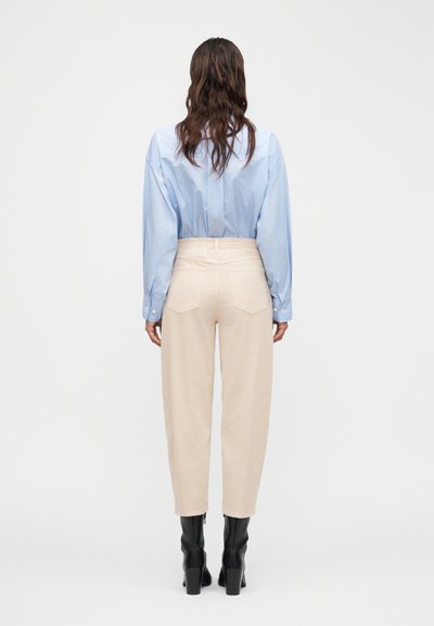 Femme aux longs cheveux bruns, portant une chemise bleu clair, un pantalon beige taille haute et des bottes à talons noires, debout devant un fond blanc.