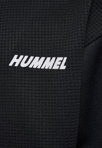 Close-up af sort tekstureret stof med hvidt HUMMEL-mærkelogo trykt på.