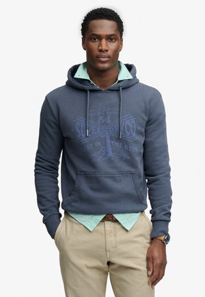 Jeune homme portant un sweat à capuche bleu par-dessus une chemise verte et un pantalon beige, debout les mains dans les poches devant un fond uni.