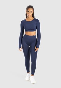 Kobieta stojąca przodem w granatowej, długiej bluzce crop top i legginsach z wysokim stanem, w białych sneakersach na jednolitym tle.