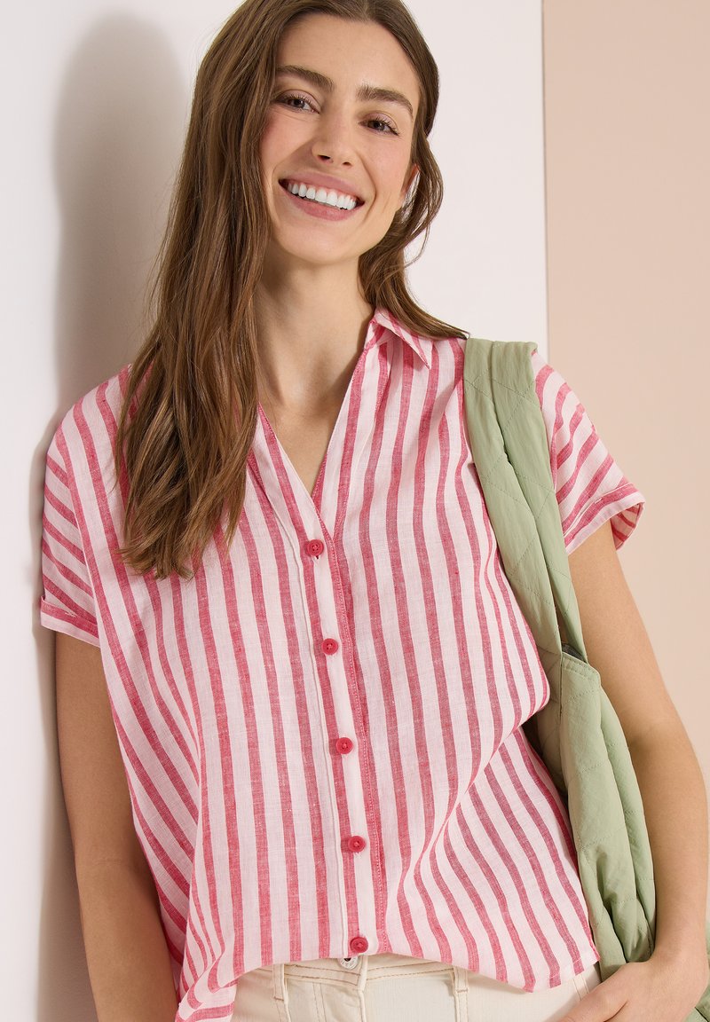 Jeune femme souriante, portant une chemise à manches courtes rayée verticalement rose et blanche, un pantalon beige, et un sac matelassé vert clair.