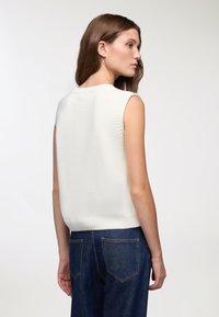 Biały bezrękawnik z dzianiny z żebrowanym dekoltem, z prostym tyłem i miękką teksturą, w połączeniu z ciemnoniebieskimi dżinsami.