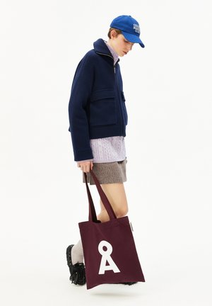 Bordeaux totebag met een witte grafische letterontwerp, gedragen met een marineblauwe jas, paarse trui en een beige rok, gecombineerd met chunkyschoenen.