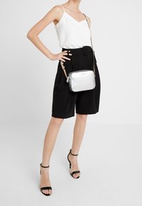 Borsa a tracolla argentata con superficie strutturata, chiusura a zip e dettaglio logo. Indossata con una cintura nera e shorts a vita alta.