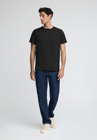 Zwarte katoenen t-shirt met korte mouwen, ronde halslijn, gecombineerd met blauwe denim jeans en lichte sneakers, staand op een effen achtergrond.