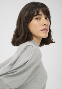 Pull en tricot gris clair avec des manches bouffantes et un col rond, présentant une texture douce. Le modèle est montré en profil.