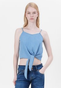 Top corto in denim azzurro chiaro con davanti annodato, spalline sottili e orlo diritto. Texture liscia con dettagli di cucitura visibili.
