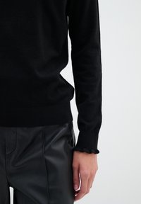 Svart stickad tröja med långa ärmar, med en vågig kant vid ärmsluten. Kombinerad med svarta skinnbyxor. Slät textur.