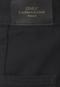 Črno denimsko blago z vidno teksturo in usnjenim etiketom, ki prikazuje "ONLY CARMAKOMA Denim" v zlatih �črkah.