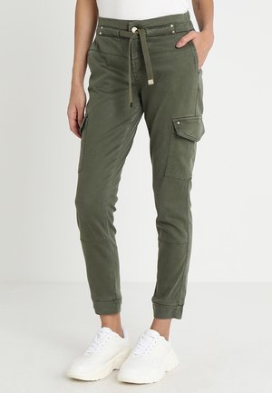 Femme portant un pantalon cargo vert olive avec taille à cordon de serrage et des baskets blanches, debout devant un fond uni clair.