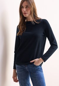 Mustahik mustahik hitam berlapis dengan lengan panjang dan leher lebar, dipadukan dengan denim biru. Kainnya tampak bertekstur dan lembut.