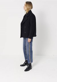 Černý dvouřadý kabát, modré úzké džíny se zlatým pruhem na boku a černé kotníkové boty. Neutrální pozadí, pohled z boku.