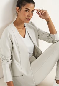 Ljust grå skräddarsydd blazer med en något strukturerad yta, kombinerad med en vit v-ringad topp och matchande slim-fit byxor.