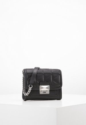 Černá prošívaná kožená crossbody taška se stříbrným řetízkovým popruhem a geometrickou stříbrnou sponou na bílém podstavci.