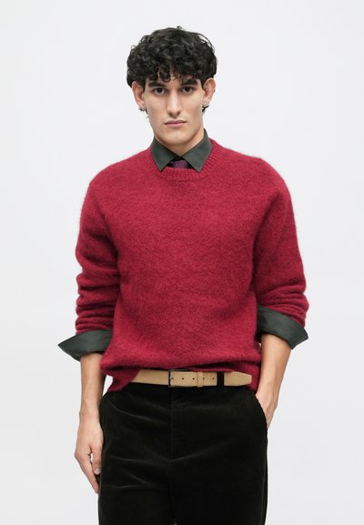 Κόκκινο φλας πουλόβερ με στρογγυλή λαιμόκοψη, σε συνδυασμό με σκούρο πράσινο πουκάμισο και παντελόνι από χοντρό πανί (κορντούρ). Διαθέτει γυρισμένα μανίκια και καφέ ζώνη. Λείο, υφασμένο ύφασμα.