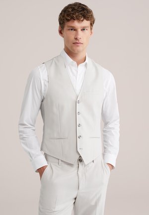 Fiatal férfi göndör hajjal, világos bézs mellényt visel fehér ing felett és hozzá illő bézs nadrágot, kezét zsebében tartja, semleges háttér előtt.