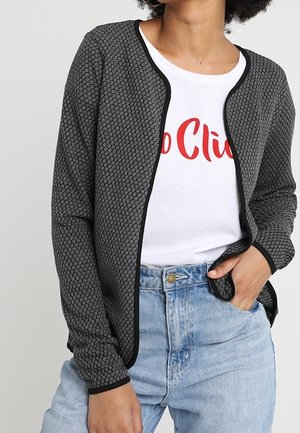 Vrouw in een grijze, gestructureerde cardigan met zwarte afwerking over een witte T-shirt met rode tekst, gecombineerd met lichtblauwe high-waisted jeans.