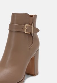 Bota de tobillo de cuero topo con tacón alto grueso, panel elástico lateral y correa decorativa con hebilla dorada.