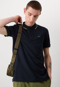 Jūros laivyno polo marškiniai su alyvuogių žalios spalvos akcentais, mygtukų uždarymu ir logotipu. Modelis neša alyvuogių žalios spalvos kryžminį krepšį ir dėvi šviesiai žalias kelnes.
