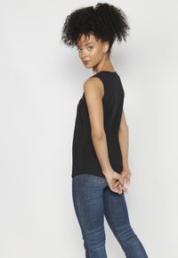 Top senza maniche nero con scollatura rotonda, caratterizzato da una texture liscia e un orlo leggermente curvo, abbinato a jeans attillati blu scuro.