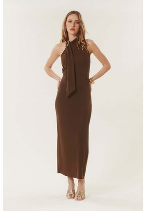 Femme debout, mains sur les hanches, portant une robe marron sans manches, longue jusqu'aux chevilles, avec un col montant et des sandales dorées sur un fond uni.
