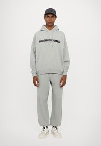 Felpa grigia in cotone con la scritta "NOON GOONS", abbinata a pantaloni della tuta coordinati. Completa il look con sneaker bianche e un berretto grigio.