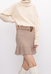 Beige Rollkragenpullover kombiniert mit einem gerippten, hellbraunen Rock mit einem Spitzenmuster und Rüsche am Saum. Als Accessoire dient ein hellbrauner Gürtel.