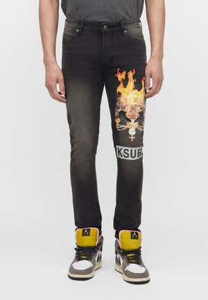 Μαύρα τζιν με γραφικό φλεγόμενου κρανίου και το κείμενο «KSUBI» στο δεξί μηρό, φορεμένα με πολύχρωμα παπούτσια υψηλού πάτου.