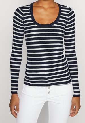 Vrouw draagt een marineblauw en wit horizontaal gestreept shirt met lange mouwen en een ronde hals, gecombineerd met witte hoge taillebroek met knopen.