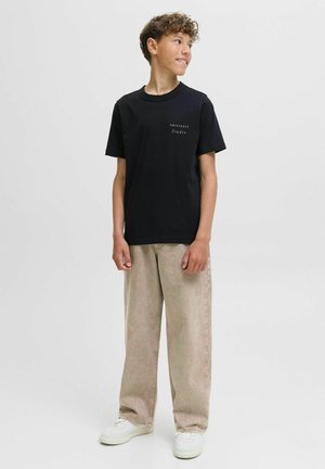 Camiseta de algodón negra con una pequeña impresión blanca de "ORIGINALS Studio". Combinada con unos vaqueros de pierna ancha en un tono beige claro y zapatillas blancas.