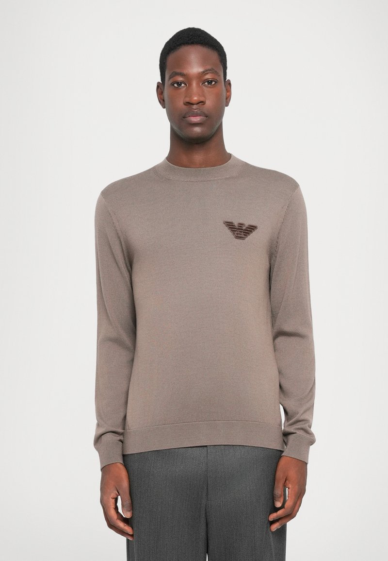 Brown knit sweater with long sleeves, round neckline, and a subtle logo in darker brown on the chest. Smooth texture, simple design.