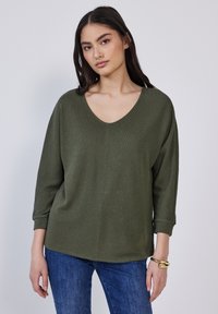 Olivgrüner Strickpullover mit weitem V-Ausschnitt, Drei-Viertel-Ärmeln und lockerer Passform, kombiniert mit blauen Jeans. Strukturiertes Material.