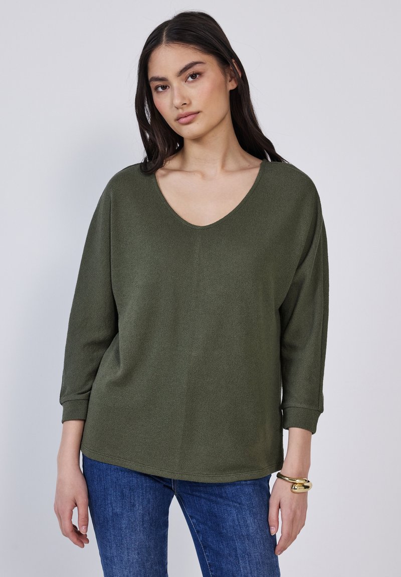 Olivgrüner Strickpullover mit weitem V-Ausschnitt, Drei-Viertel-Ärmeln und lockerer Passform, kombiniert mit blauen Jeans. Strukturiertes Material.