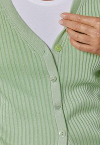 Grüne gerippte Strickjacke mit V-Ausschnitt und fünf hellgrünen Knöpfen. Über einem weißen Hemd getragen, das aus einem strukturierten Stoff mit vertikalen Streifen besteht.