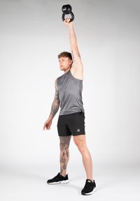 Atelet halli halli särk ja mustades lühikeses püksides tõstab musta kettlebelli pea kohal. Silmapaistvad tätoveeringud käel ja keeruline jalatsite disain.