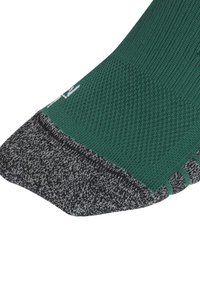 Calcetín verde con tejido texturizado, presenta una puntera y un talón en gris oscuro, y una sección de tobillo acanalada. Marca distintiva en negro y blanco en el lateral.