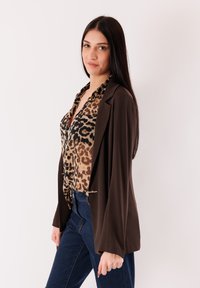 Blazer marrone a maniche lunghe sopra una camicetta con stampa leopardata. La camicetta presenta un design con bottoni. Jeans blu scuro completano l'outfit.