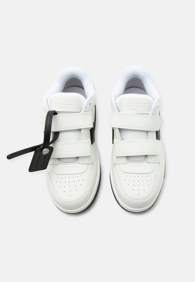 Ζευγάρι λευκά αθλητικά παπούτσια με βέλκρο και μαύρη ετικέτα στο αριστερό παπούτσι, εμφανιζόμενα από πάνω σε απλό ανοιχτό φόντο.