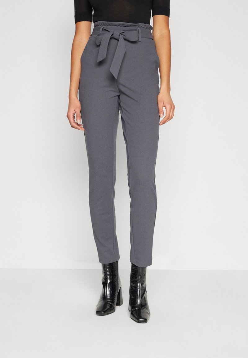 Femme portant un pantalon gris taille haute avec une ceinture nouée et des bottines noires à talons hauts sur un fond blanc uni.
